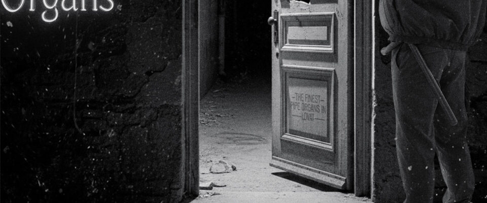 Alley door broken open and throw wide. A figure observes it from outside. A neon sign that say Barnabé Organs glows to one side.